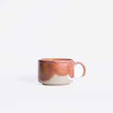 A handcrafted ceramic cup with a handle, displaying a lively organic pattern in coral red and white finishes, showcased against a pristine white background.