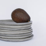 Close-up detail of handcrafted gray ceramic plates with a matte, earthy texture, an avocado resting on the top plate for scale, set against a clean white background.