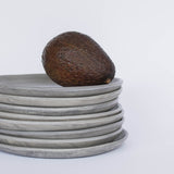 Close-up detail of handcrafted gray ceramic plates with a matte, earthy texture, an avocado resting on the top plate for scale, set against a clean white background.