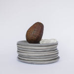 Pile of handcrafted gray ceramic plates with a matte, earthy texture, an avocado resting on the top plate for scale, set against a clean white background.