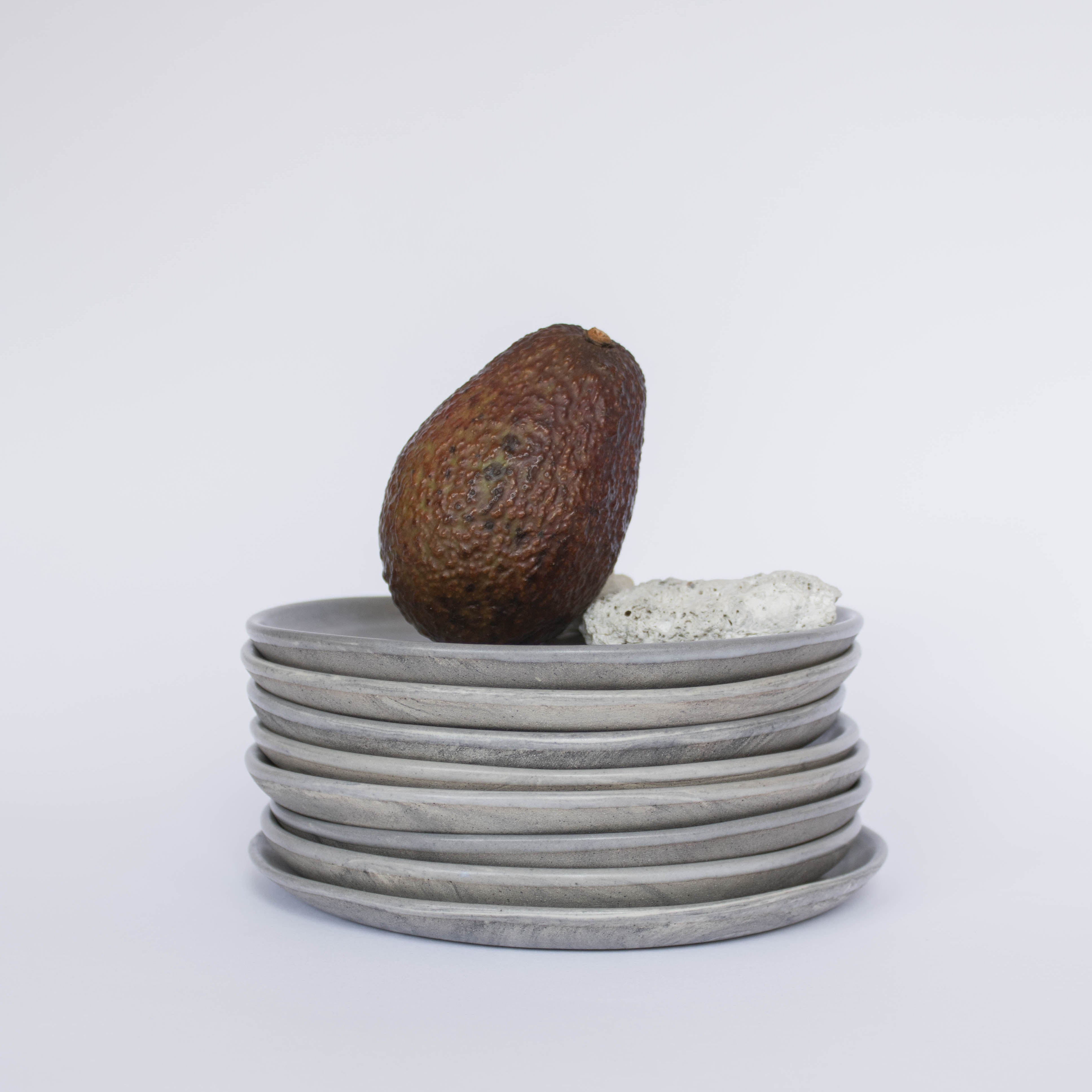 Pile of handcrafted gray ceramic plates with a matte, earthy texture, an avocado resting on the top plate for scale, set against a clean white background.