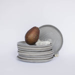 Pile of handcrafted gray ceramic plates with a matte, earthy texture, an avocado resting on the top plate for scale, set against a clean white background.