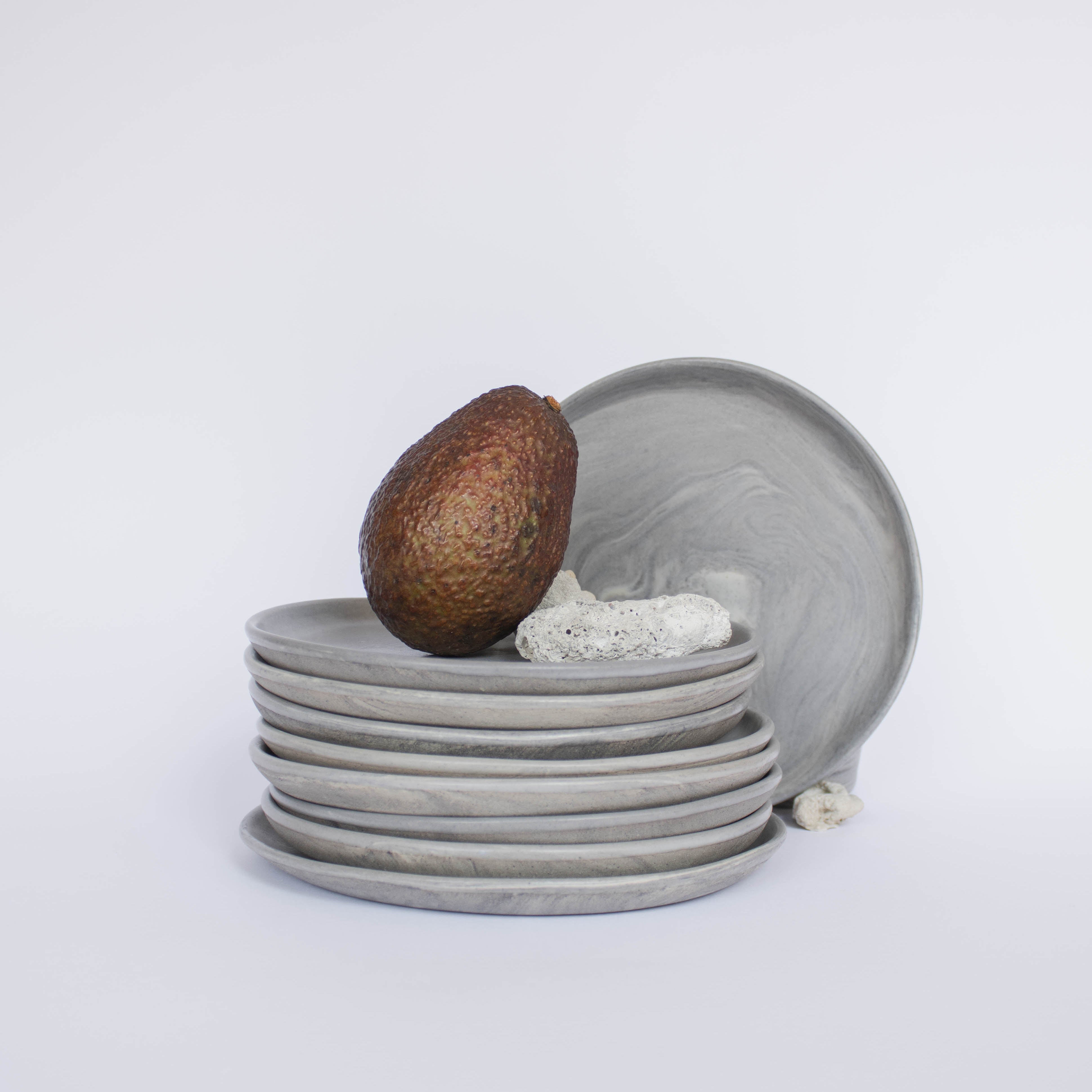 Pile of handcrafted gray ceramic plates with a matte, earthy texture, an avocado resting on the top plate for scale, set against a clean white background.