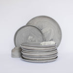 Pile of handcrafted gray ceramic plates with a matte, earthy texture, with two representative plates set vertically behind, all against a clean white background.