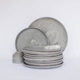 Pile of handcrafted gray ceramic plates with a matte, earthy texture, with two representative plates set vertically behind, all against a clean white background.