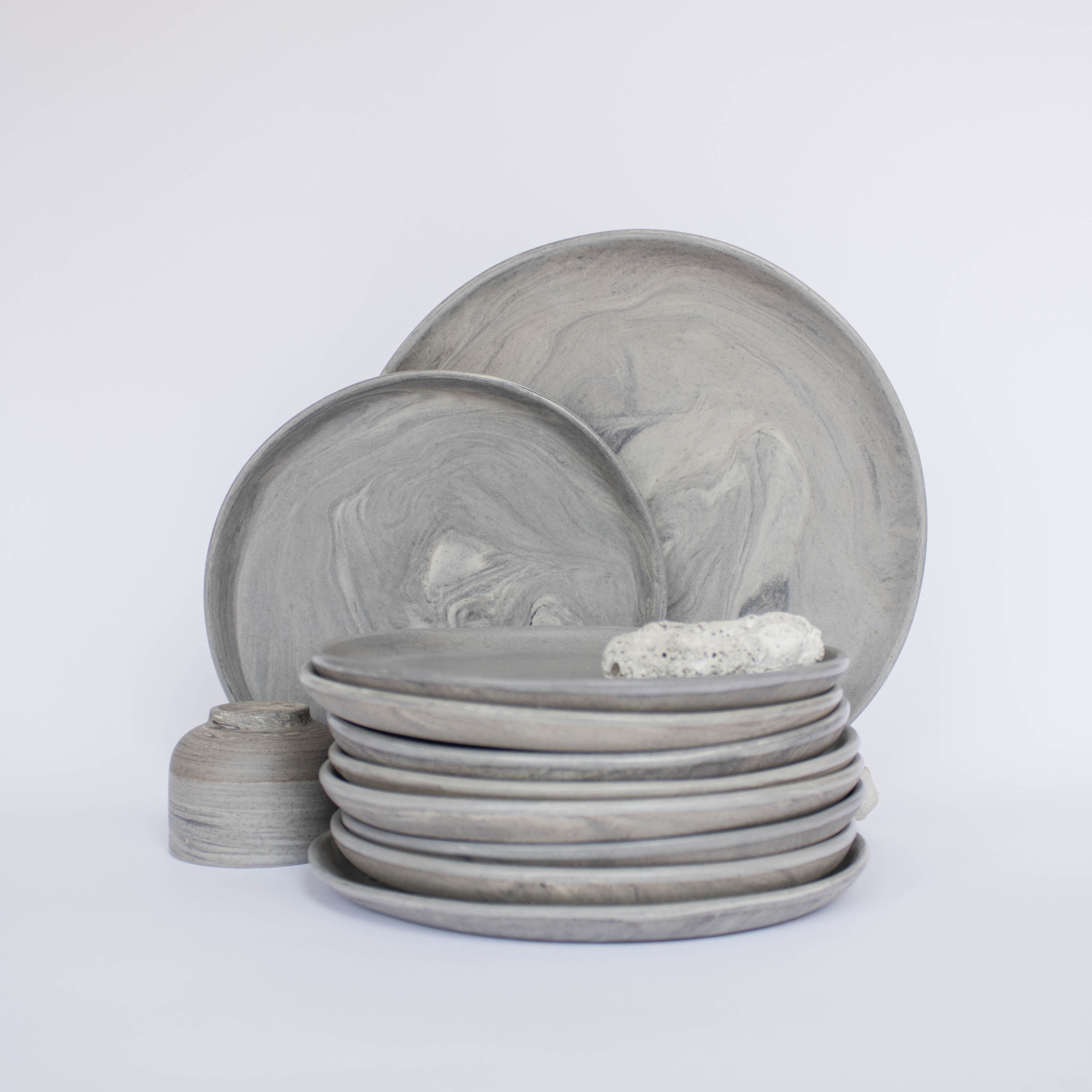 Pile of handcrafted gray ceramic plates with a matte, earthy texture, with two representative plates set vertically behind, all against a clean white background.