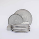 Pile of handcrafted gray ceramic plates with a matte, earthy texture, with two representative plates set vertically behind, all against a clean white background.