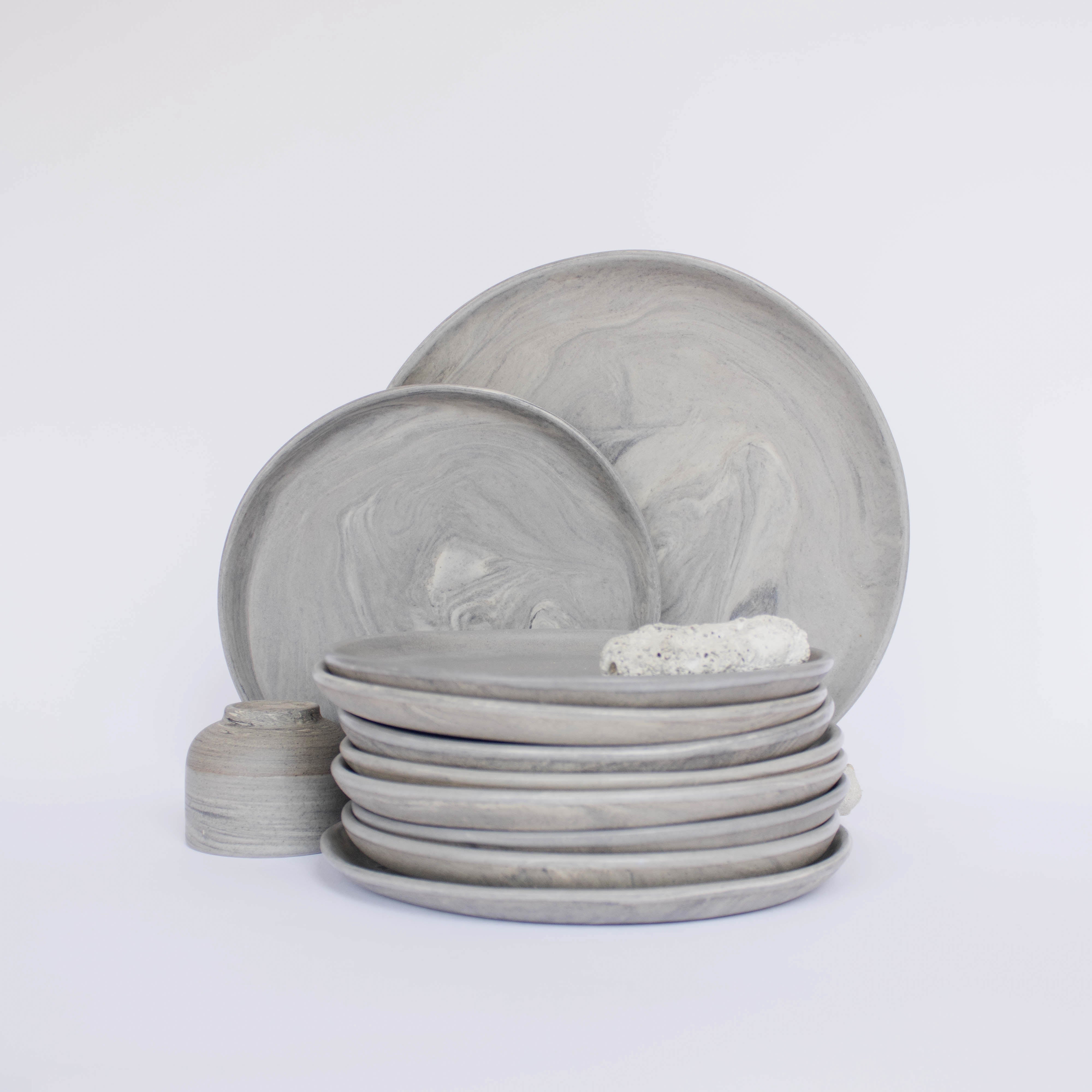 Pile of handcrafted gray ceramic plates with a matte, earthy texture, with two representative plates set vertically behind, all against a clean white background.