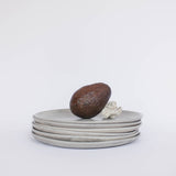 Pile of handcrafted gray ceramic plates with a matte, earthy texture, an avocado resting on the top plate for scale, set against a clean white background.