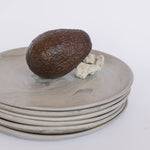 Pile of handcrafted gray ceramic plates with a matte, earthy texture, an avocado resting on the top plate for scale, set against a clean white background.