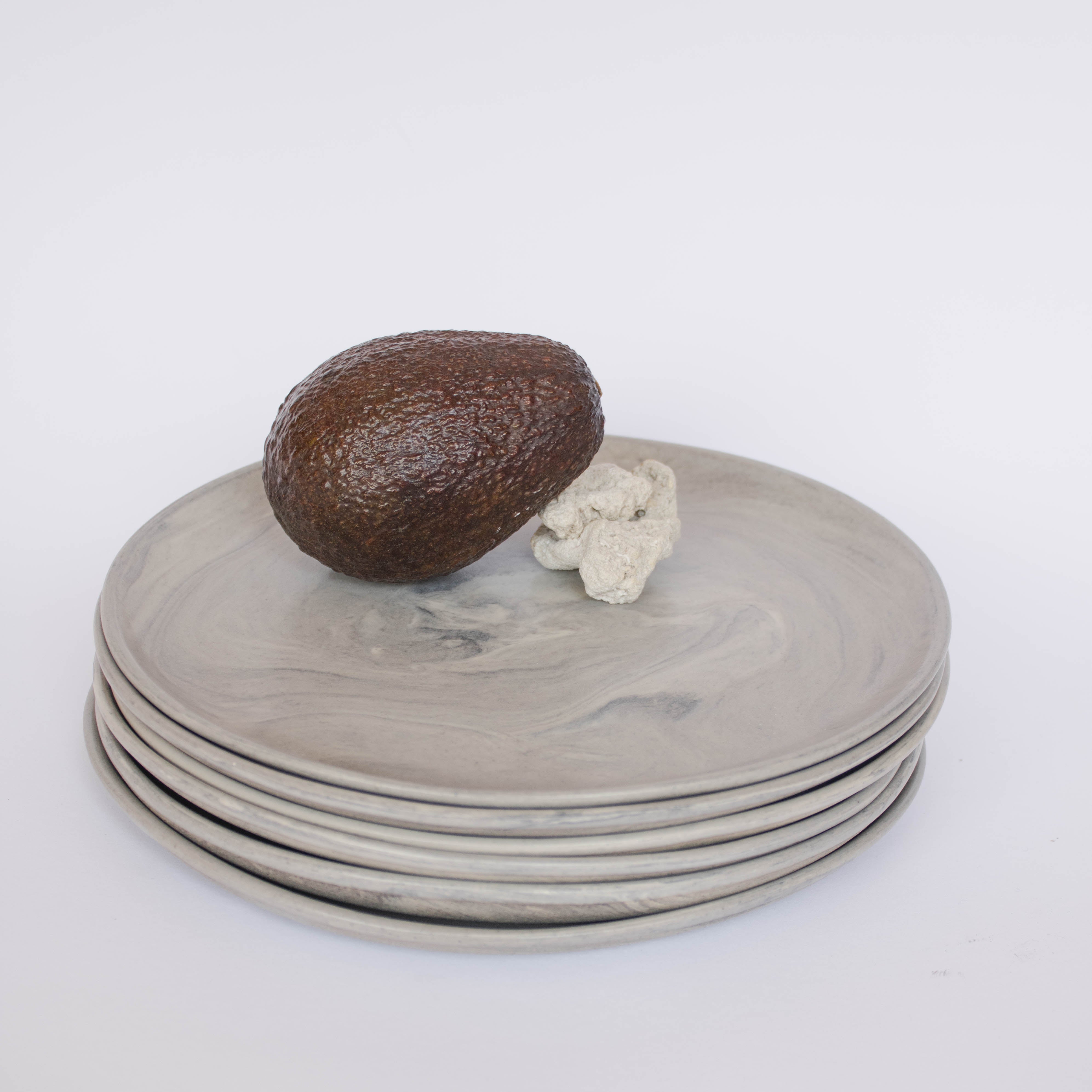 Pile of handcrafted gray ceramic plates with a matte, earthy texture, an avocado resting on the top plate for scale, set against a clean white background.