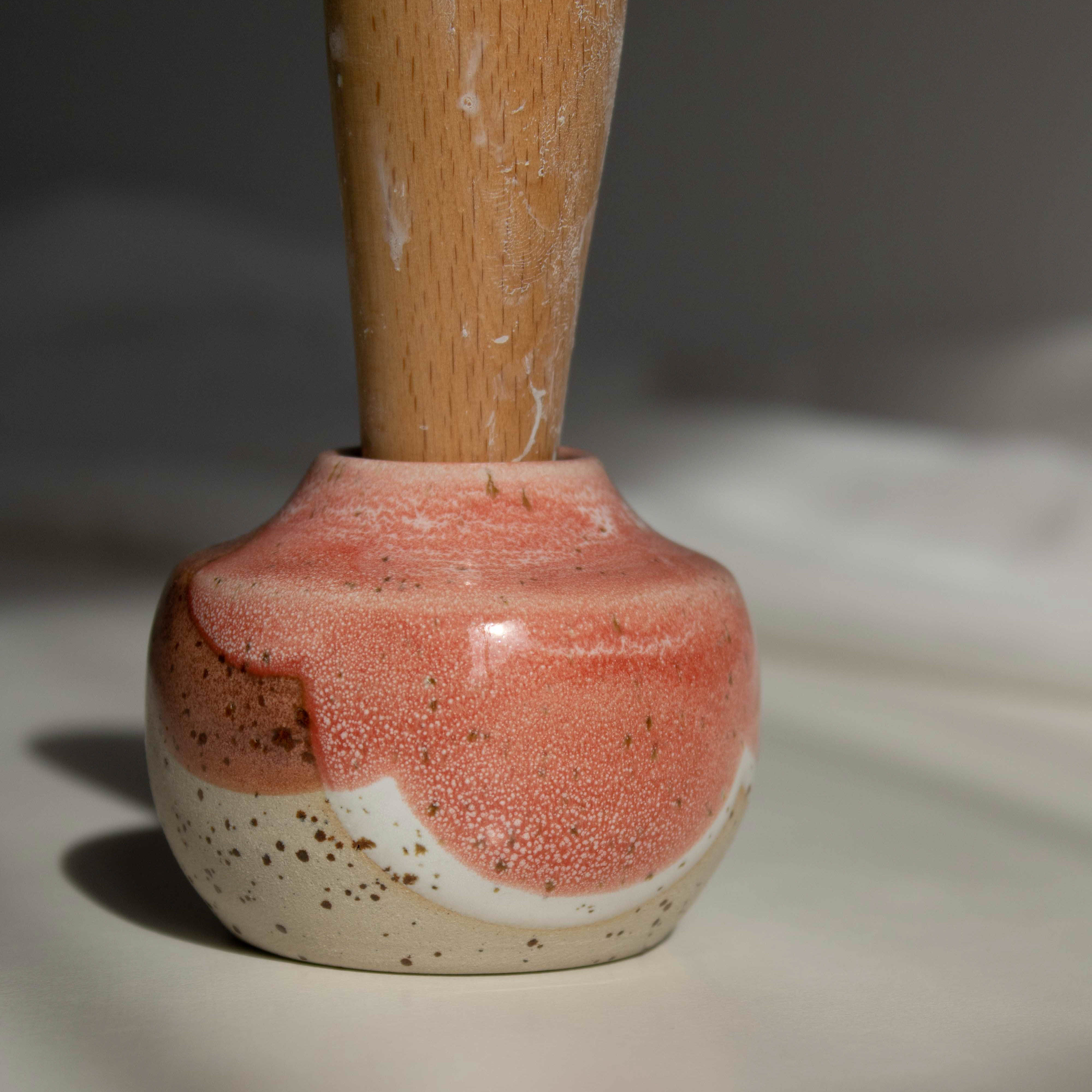 Close-up of a handcrafted miniature ceramic vase in coral red finish, featuring a maker's mark stamp with a wooden handle, photographed in natural daylight.
