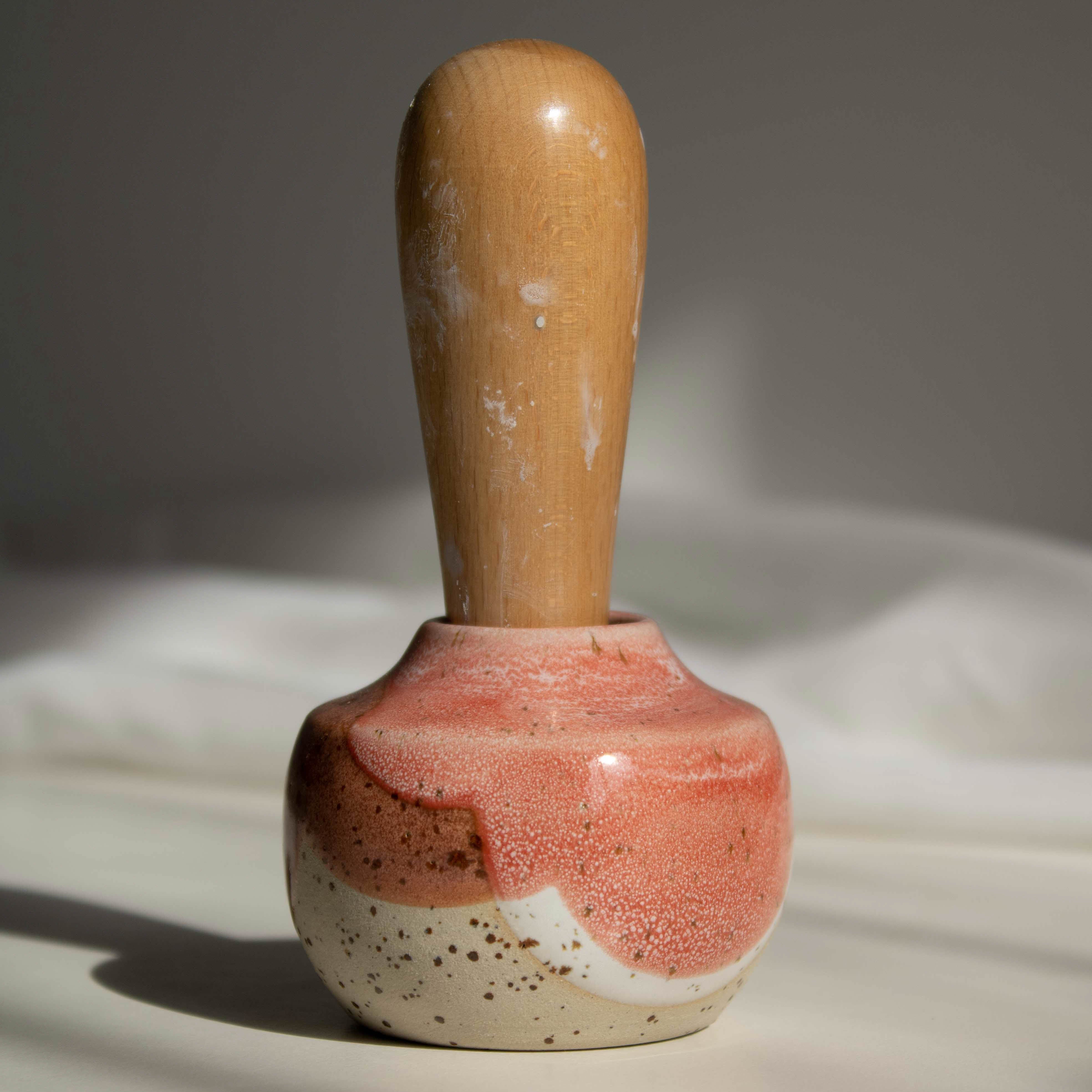 Close-up of a handcrafted miniature ceramic vase in coral red finish, featuring a maker's mark stamp with a wooden handle, photographed in natural daylight.