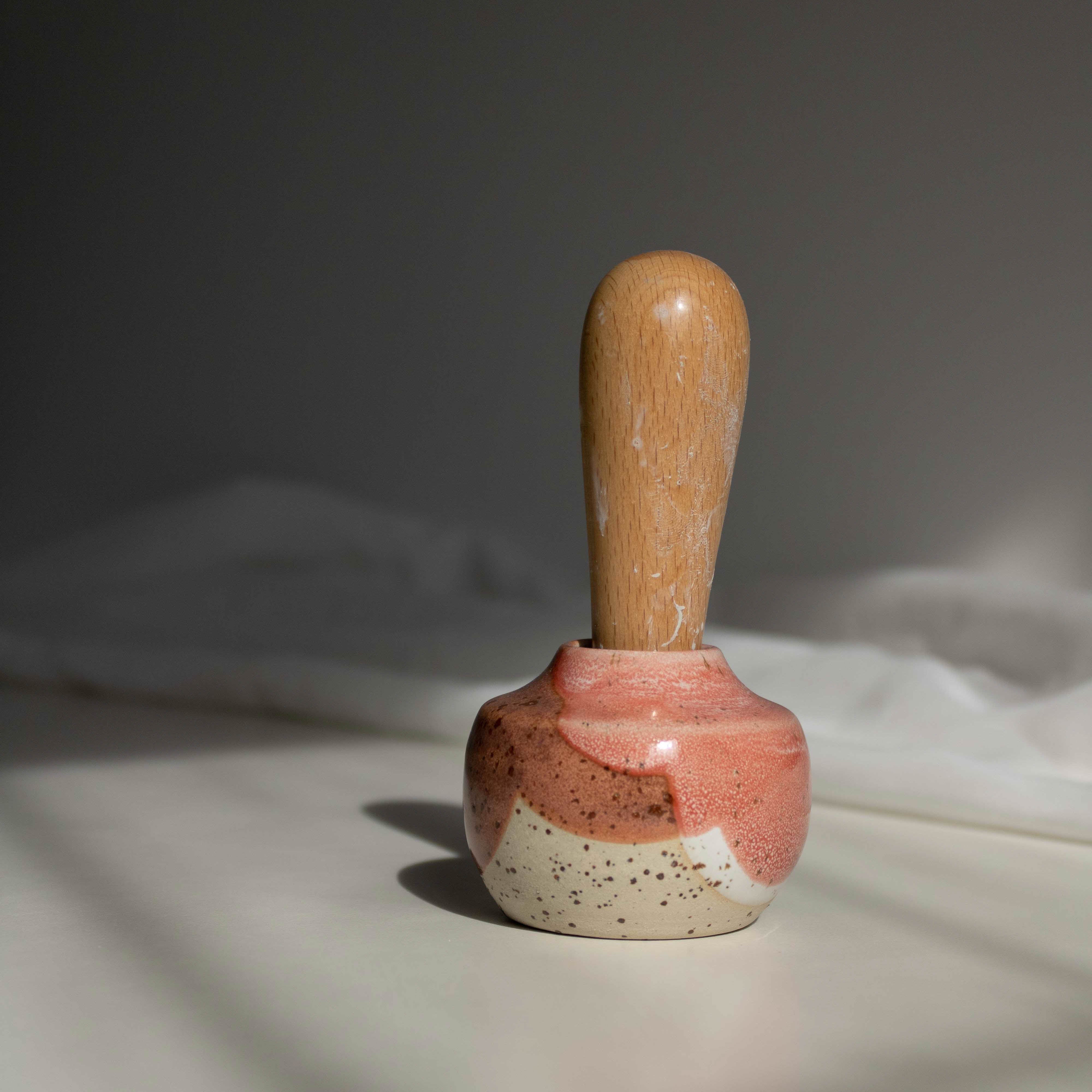Handcrafted miniature ceramic vase in coral red finish, featuring a maker's mark stamp with a wooden handle, photographed in natural daylight.