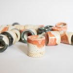 Foregrounded handmade ceramic toothbrush or pen holder in a coral red finish, against a backdrop of blurred holders scattered in the background.