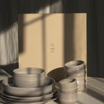 A set of matte ceramic tableware in warm off-white and light beige tones, including stacked plates, bowls, and small cups, arranged in front of a large beige gift box on a white surface with dramatic window shadow lighting.