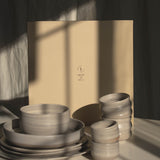 A set of matte ceramic tableware in warm off-white and light beige tones, including stacked plates, bowls, and small cups, arranged in front of a large beige gift box on a white surface with dramatic window shadow lighting.