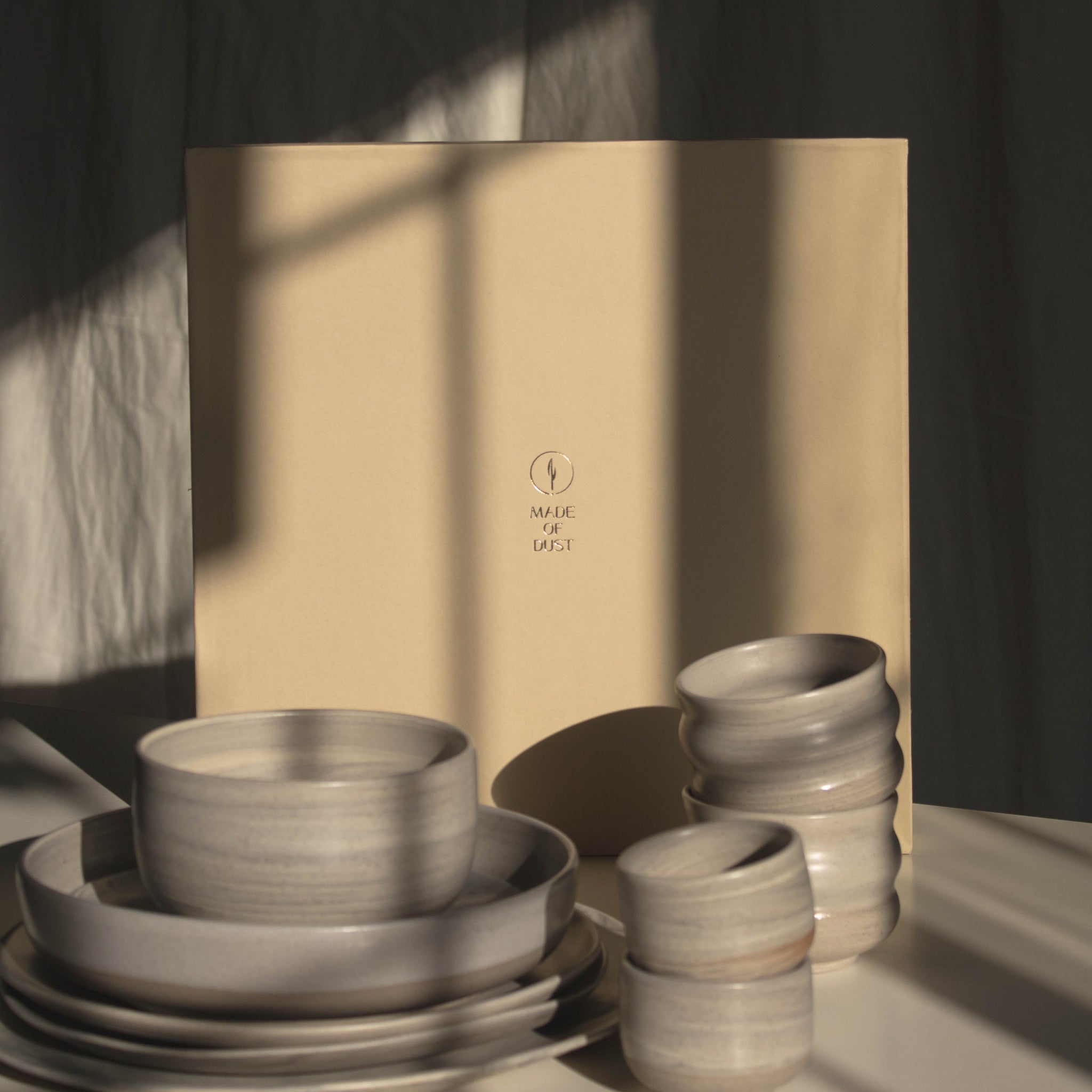 A set of matte ceramic tableware in warm off-white and light beige tones, including stacked plates, bowls, and small cups, arranged in front of a large beige gift box on a white surface with dramatic window shadow lighting.
