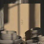 A set of matte ceramic tableware in a warm gray tone, including bowls, plates, and cups, arranged in front of a large warm beige paper box with a small brand logo.