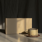 A beige ceramic coffee mug on a matching saucer sits beside a rectangular beige gift box on a tabletop, with dramatic sunlight casting shadows against a dark backdrop.