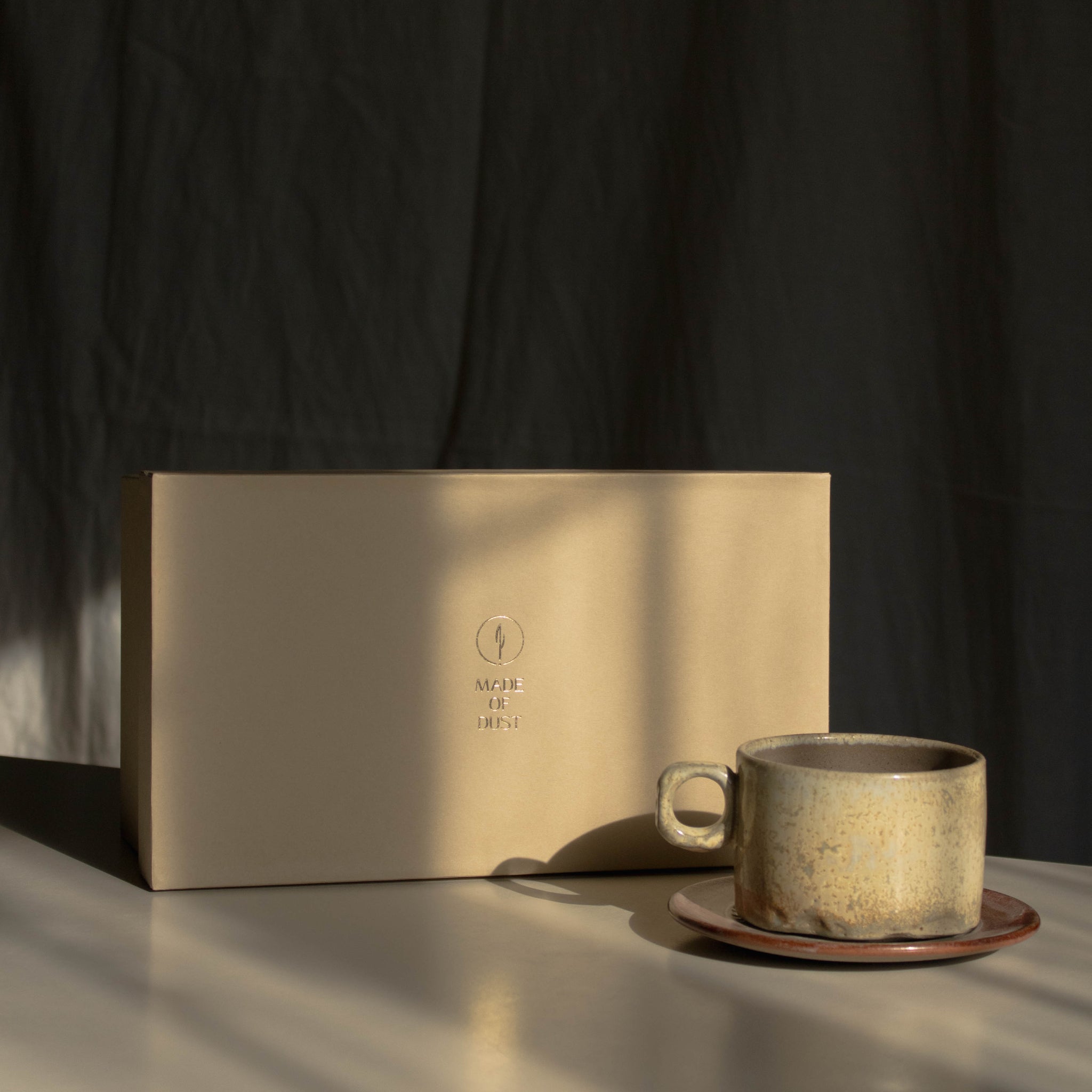 A beige ceramic coffee mug on a matching saucer sits beside a rectangular beige gift box on a tabletop, with dramatic sunlight casting shadows against a dark backdrop.