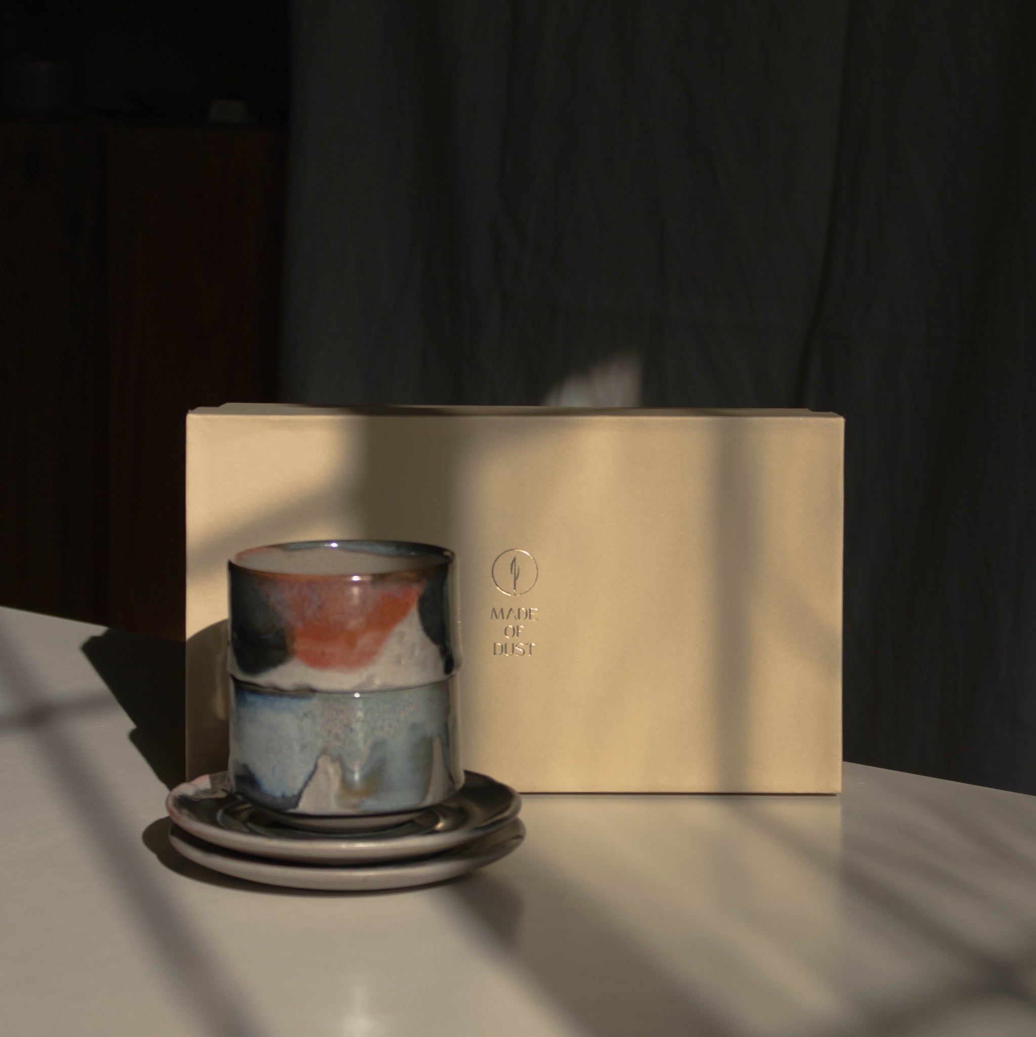 A stack of two ceramic cups on two matching saucers beside a cream-colored Special Occasion Gift Box, lit by dramatic sunlight and strong shadows.