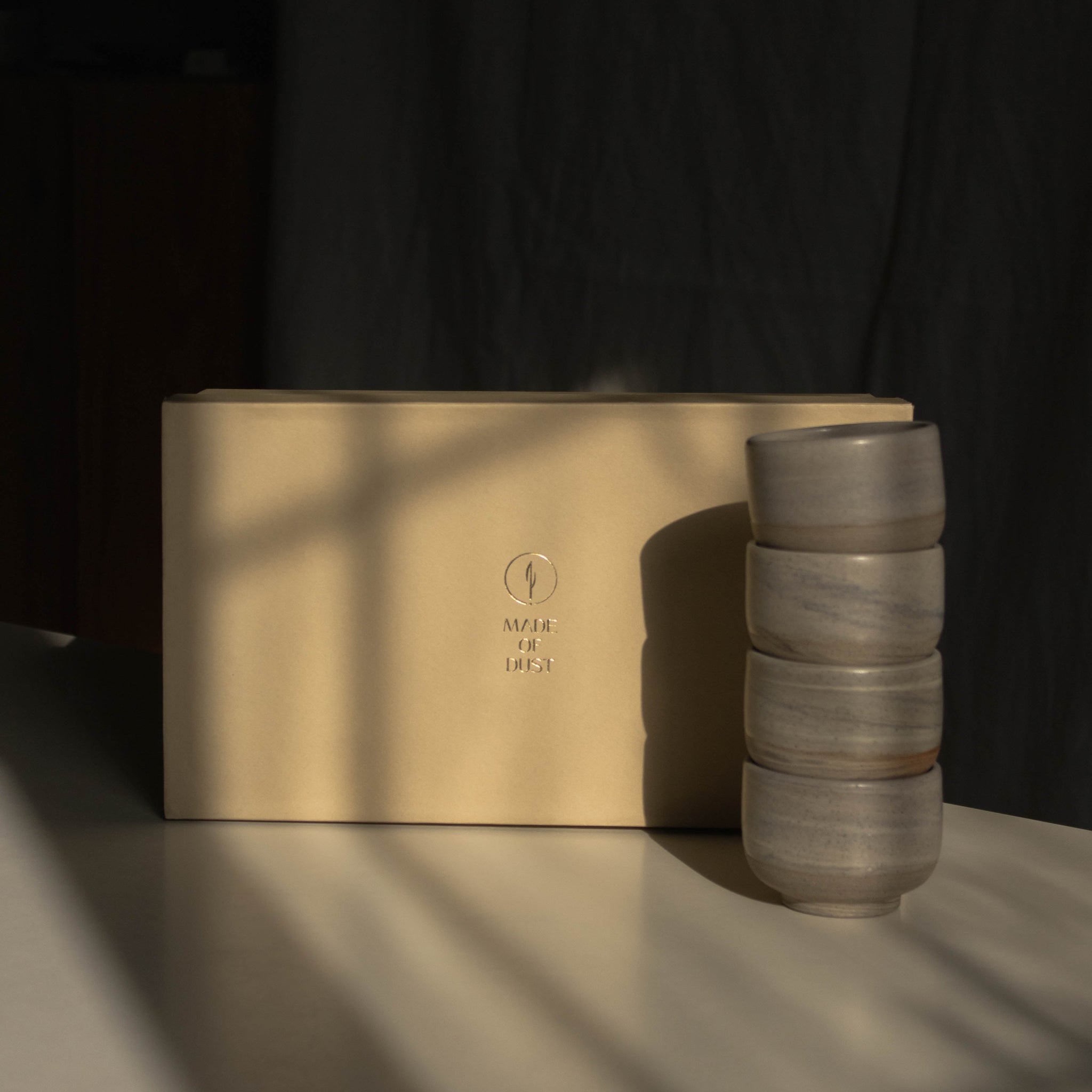 Warm beige Special Occasion Gift Box with a gold Made of Dust logo beside four stacked marbled gray ceramic cups in dramatic window light.