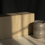 A warm beige rectangular Special Occasion Gift Box with the Made of Dust logo sits on a tabletop beside two Ripple ceramic cups, in dramatic sunlight with a dark draped background.
