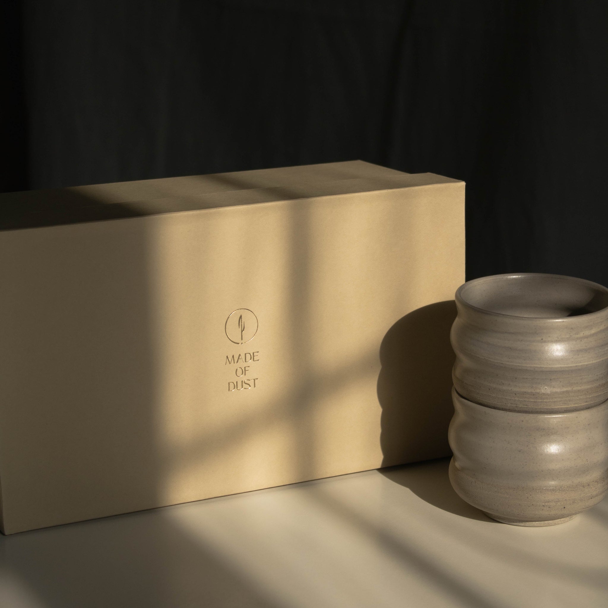 A warm beige rectangular Special Occasion Gift Box with the Made of Dust logo sits on a tabletop beside two Ripple ceramic cups, in dramatic sunlight with a dark draped background.