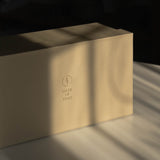 A minimalist warm beige gift box with a gold Made of Dust logo, resting on a surface in dramatic diagonal sunlight and shadows.