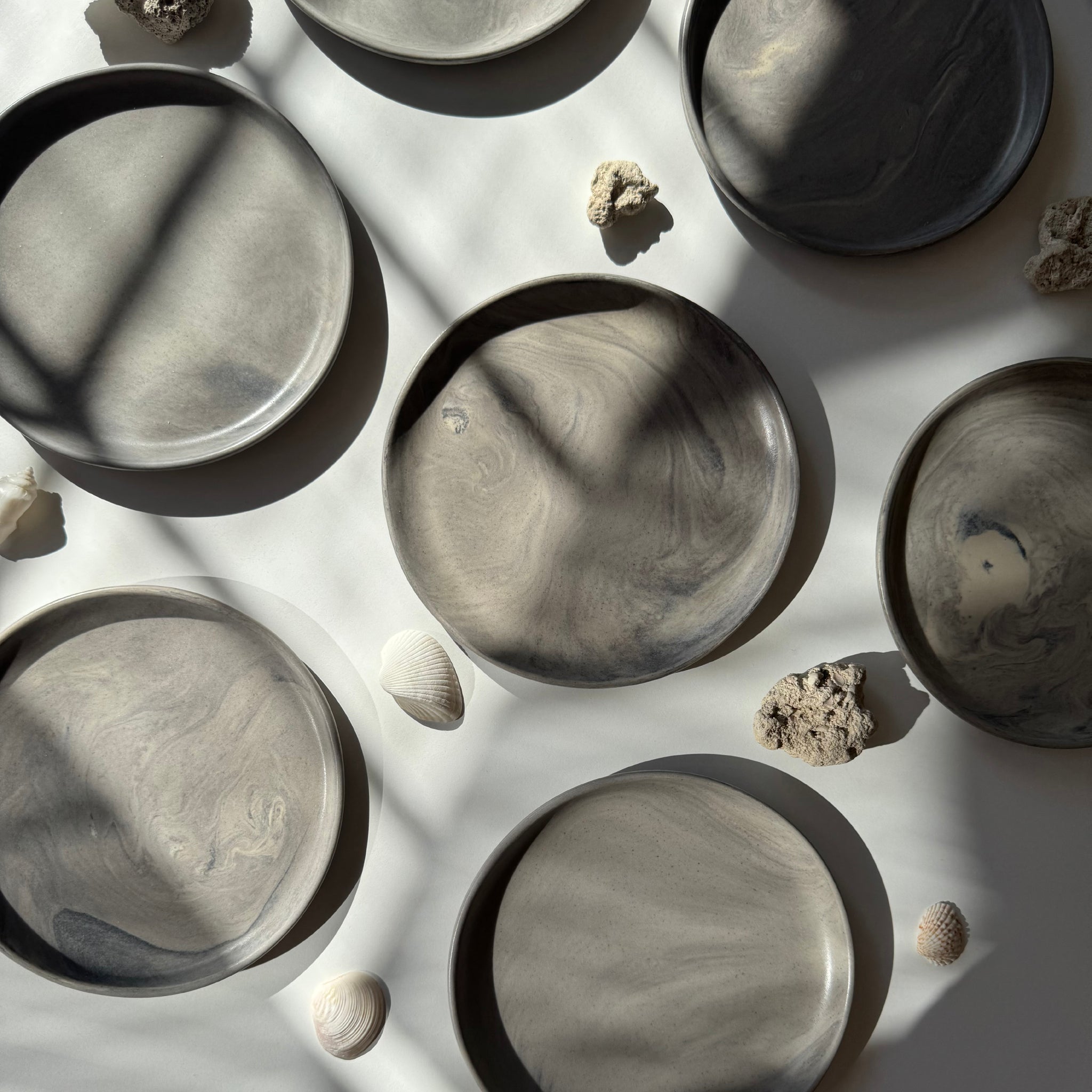 Overhead view of handmade ceramic plates with a matte marble finish, arranged on a white surface where sunlight creates dramatic shadow play.