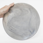 A handmade ceramic plate in gray with a matte marble finish, held against a white background.
