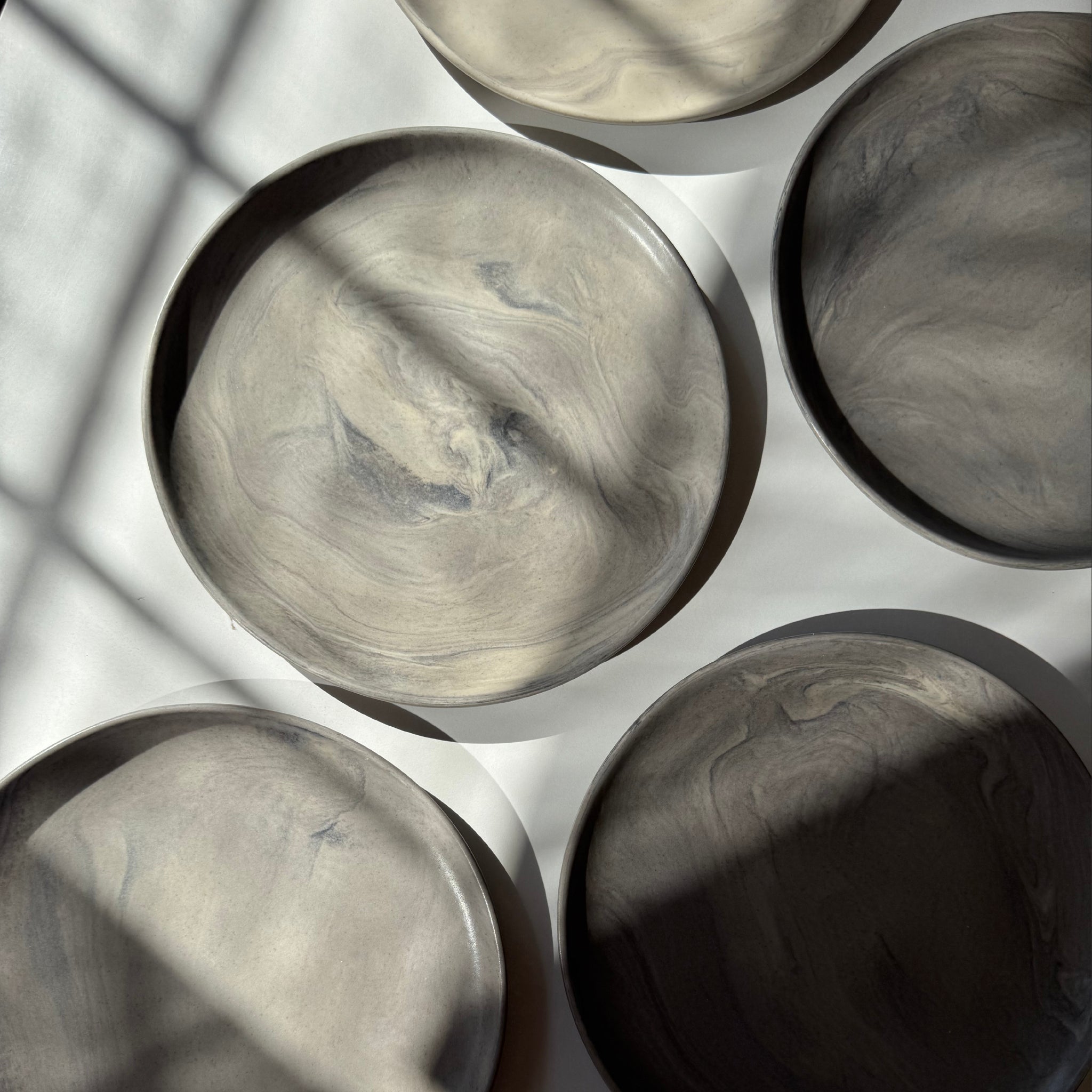 Overhead view of handmade ceramic plates with a matte marble finish, arranged on a white surface where sunlight creates dramatic shadow play.