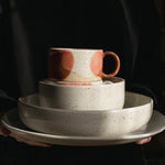 Hands holding handcrafted speckled white plates, a bowl, and a pink cup against a black backdrop, bathed in sunlight.