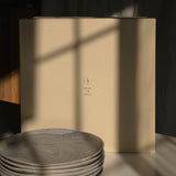 A stack of round gray marbled plates sits in front of a tall beige box with a small embossed logo, photographed in dramatic sunlight with window-frame shadows across the scene.