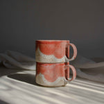 Two coral red cups with handles illuminated by a beam of light.